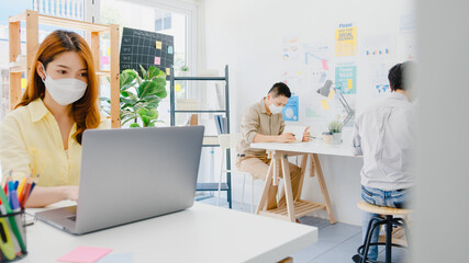 Asia female wear face mask for social distancing in new normal situation for virus prevention while using laptop and separated by acrylic partition stand in office. Life and work after corona virus.