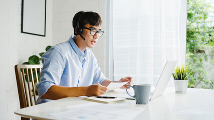 Young Asia businessman using laptop talk to colleagues about plan in video call while smart working from home at living room. Self-isolation, social distancing, quarantine for corona virus prevention.