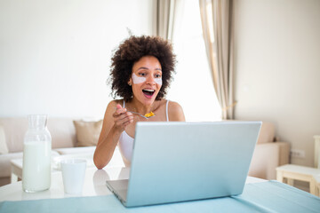 Image of happy young amazing woman sitting indoors at the table with laptop holding corn flakes. Looking at laptop computer and talking to her friends via video call.