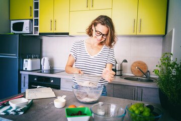 Beautiful young woman preparing dessert at home in the kitchen and sifting flour, hobby and entertainment, food concept