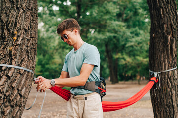Stylish young man ties a hammock in the woods to a tree, ties the slings with his hands. The...
