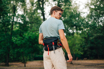 Handsome young man in summer casual clothes posing in the woods with a hiking bag on his belt, looking away. Hipster man with waist bag in the park.