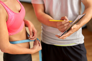 fitness, sport and people concept - close up of young woman with measuring tape and personal trainer with clipboard in gym
