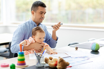 remote job, multi-tasking and family concept - middle-aged father with baby using voice command recorder on smartphone at home office