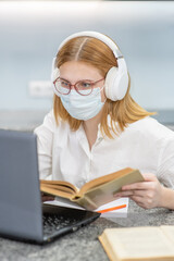 Teen girl wearing medical protective mask and headphones is engaged in distance learning with a laptop at home. Quarantine and coronavirus epidemic concept