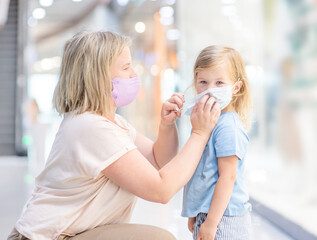 Woman helps put on a mask for her little daughter while they are in a public crowded place - in a shopping mall or airport