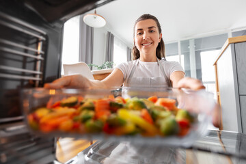 culinary, food and people concept - woman cooking vegetables in baking dish in oven at home kitchen