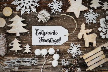 One White Label With German Text Entspannte Feiertage Means Merry Christmas. Frame Of Christmas Decoration Like Tree, Sled, Star And Fir Cone. Wooden Background With Snowflakes