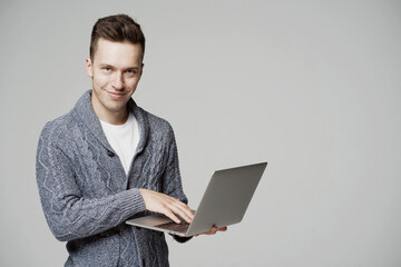 smart young man student of European appearance, working on a laptop and looking at the camera, smiling. stylish brunette hairstyle. he's wearing a gray sweatshirt. gray background