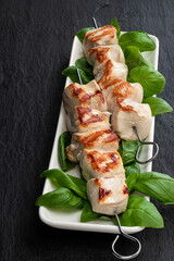 Grilled chicken breast fillet with basil on a skewers on black stone background