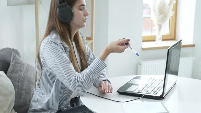 Serious girl student wears headphones, studies with internet teacher, learns language. focused young woman talks to tutor, looks at laptop, writes notes. online education, video call tutoring concept