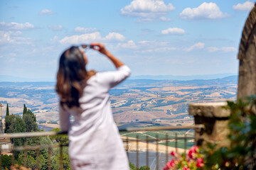 Fototapeta na wymiar Holidays in Tuscany. Middle aged woman with long hair wearing a white dress watches the landscape of Tuscany from the terrace of a medieval house, enjoying wine. Montalcino, Italy.