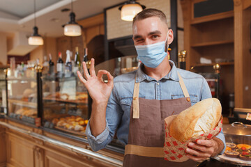 Male baker wearing medical face mask, selling bread during coronavirus pandemic quarantine, showing...