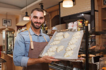 Cheerful male baker holding tray of raw croissants ready for baking