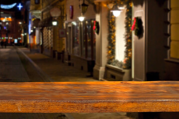 Fototapeta premium Old shabby board on a background of blurred Christmas market. Brown table top in sharpness. Christmas decorations, lights and lanterns in the background. Layout for design. Free space for products.