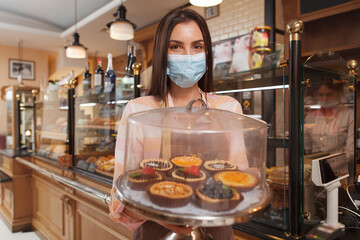 Female baker wearing medical face mask, carrying tray with desserts working at her bakery during covid-19 pandemic quarantine