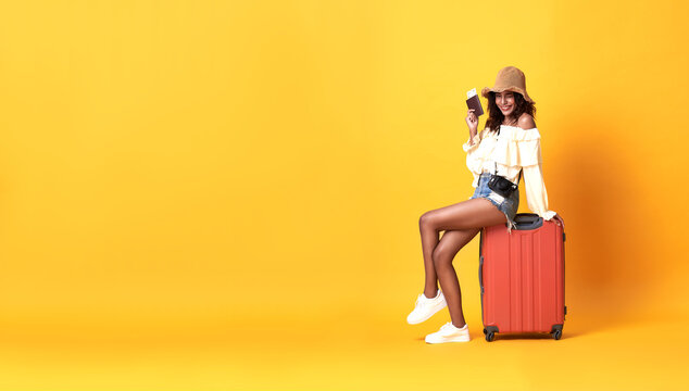 Cheerful young african woman dressed in summer clothes sitting on a suitcase on copy space yellow background.