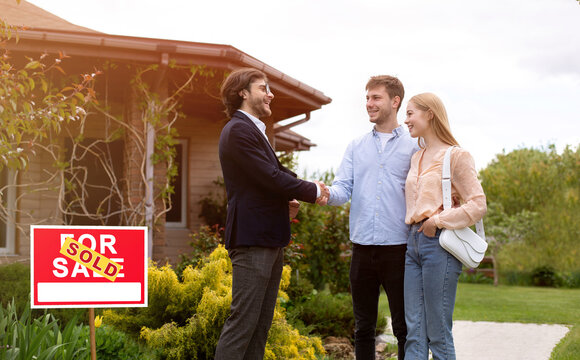 Cheerful realtor shaking hands with clients in front of residential building outdoors, free space