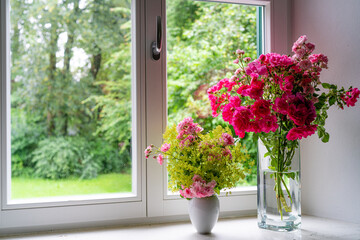 Rosenstrauß mit roten Rosen in Glasvase, auf Fensterbrett mit Blick hinaus in einen weiten Garten