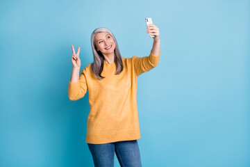 Photo of positive old woman travel trip take selfie smartphone make v-sign wear jumper denim jeans isolated over blue color background