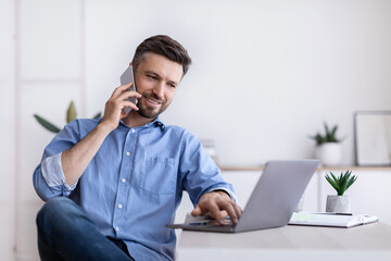 Cheerful Young Man Working Remotely From Home Office With Cellphone And Laptop