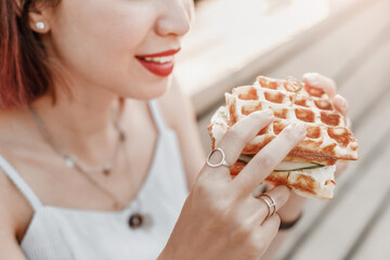 Smiling hipster woman eating a delicious sandwich with a loaf of a checkered Viennese waffle. Modern Street food concept