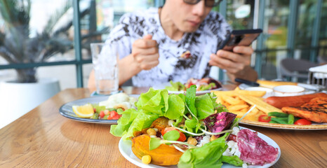 Selective focus of  green salad  plate and  Asian Man using  as looking at his smartphone with having foods of lunch in the restaurant Background