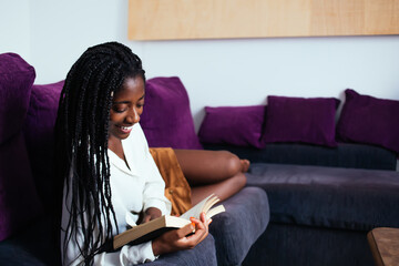 Cheerful dark skinned woman with braids smiling while read novel on free time in comfortable living room, pretty african american hipster girl lying on sofa enjoying educational literature on leisure