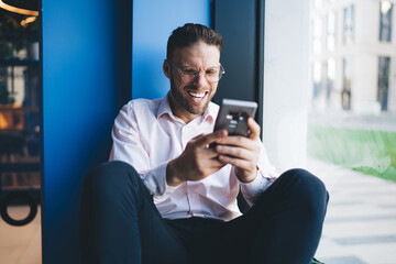 Cheerful man laughing while using smartphone in modern workspace