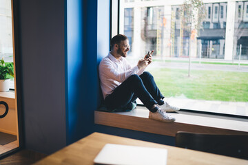 Cheerful young man using smartphone near window in modern workspace