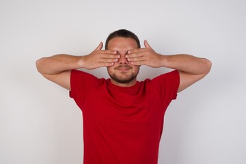 Young caucasian man wearing red t-shirt over white background covering eyes with hands smiling cheerful and funny. Blind concept.