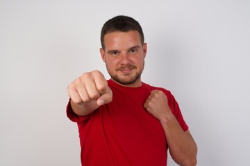 Young caucasian man wearing red t-shirt over white background Punching fist to fight, aggressive and angry attack, threat and violence