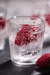 Alcohol shots of berries cocktail with a raspberry on black stone tray. Glass of sparkling water on wooden table
