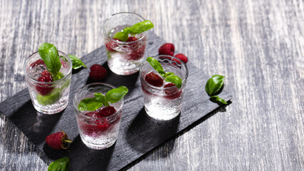Alcohol shots of berries cocktail with a raspberry and basil on black stone tray. Glass of sparkling water on wooden table
