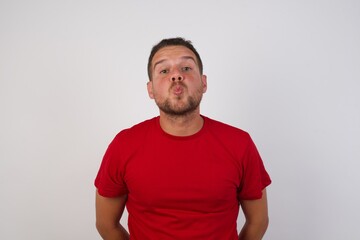 Shot of pleasant looking Young handsome Cucasian man wearing red shirt standing against white background pouts lips, looks with green eyes at camera,  has fun with girlfriend, Human facial expressions