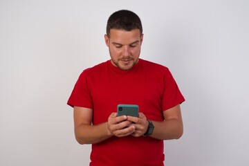Portrait of nice  Young handsome Cucasian man wearing red shirt standing against white background using mobile phone chatting free time .