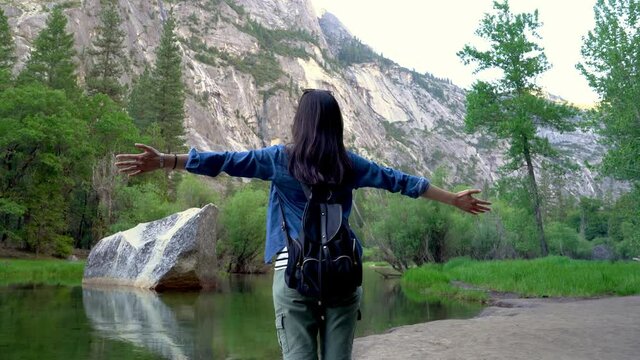 Smiling young asian woman stretching hands feeling free standing by mirror lake. girl backpacker with arms wide open outdoors on sunny summer day in forest yosemite national park california usa.