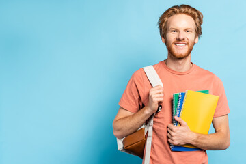 redhead student holding notebooks and touching backpack on blue