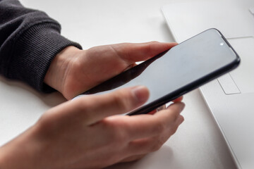 European child hands holding black smartphone with fingers and black screen on white desk infront of white notebook at school for research and mobile gaming or game addiction of kids and grown ups