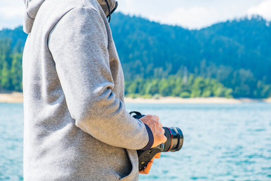 Anonymous man in hoodie holding DSLR camera in hands next to lake in nature, no face, half body, landscape photography shooting concept
