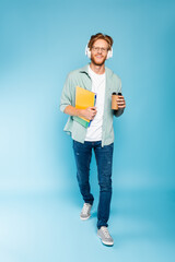 bearded student in glasses and wireless headphones holding paper cup and notebooks while walking on blue