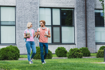 Students holding paper cups, books and digital tablet while walking on grass near campus