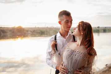 A beautiful wedding couple on the riverbank at sunset. A woman in a gray dress with flowers, a man in a suit and suspenders hug each other, love. Lovers on a walk