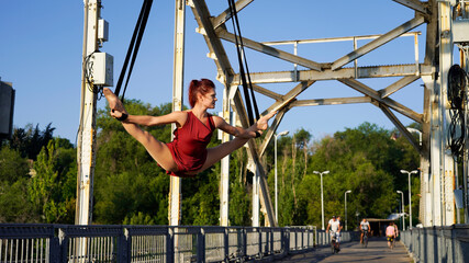 Circus performer on aerial straps performs an aerial act at dawn. flexible, athletic, aerial gymnast in city on metal trusses of bridge engaged in aerial gymnastics. urban acrobatic street show. split