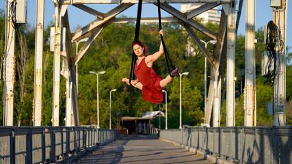 Obraz premium Beautiful sports girl aerialist. circus acrobat on aerial straps, metallic trusses of the bridge. flexible gymnast performs splits against the sky, street show