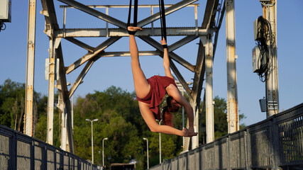 Beautiful young woman gymnast hanging in a flexible pose on an iron bridge in the rays of sunlight Professional circus performer, graceful flying on aerial straps