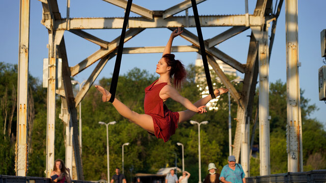Beautiful sports girl aerialist. circus acrobat on aerial straps, metallic trusses of the bridge. flexible gymnast performs splits against the sky, street show