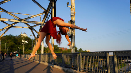Beautiful young woman gymnast hanging in a flexible pose on an iron bridge in the rays of sunlight Professional circus performer, graceful flying on aerial straps