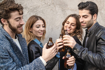 Fashionable group of best friends clinking with beers bottles against a concrete wall. Millennial generation trendy people sharing positive emotions having fun at outdoors party together