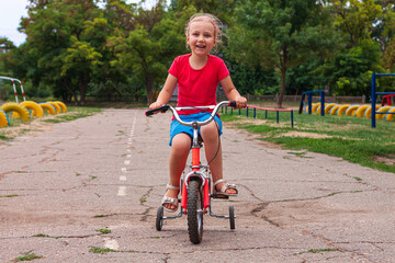 Child riding on bicycle. Blonde smiling girl on red bike on asphalt road in summer. Active...
